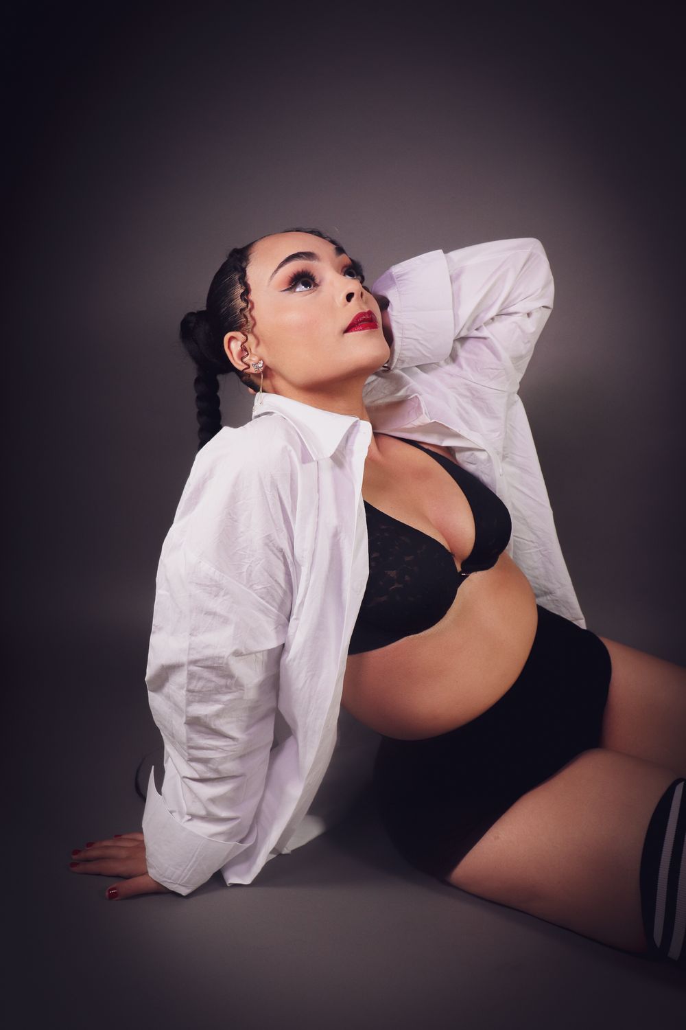 Woman in black lingerie and open white shirt posing on floor against dark studio background.