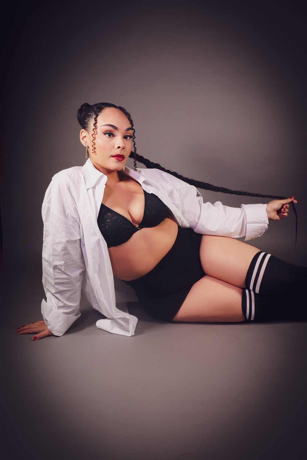 Woman in black lingerie and white shirt poses on studio floor with long braided hair and thigh-high socks.