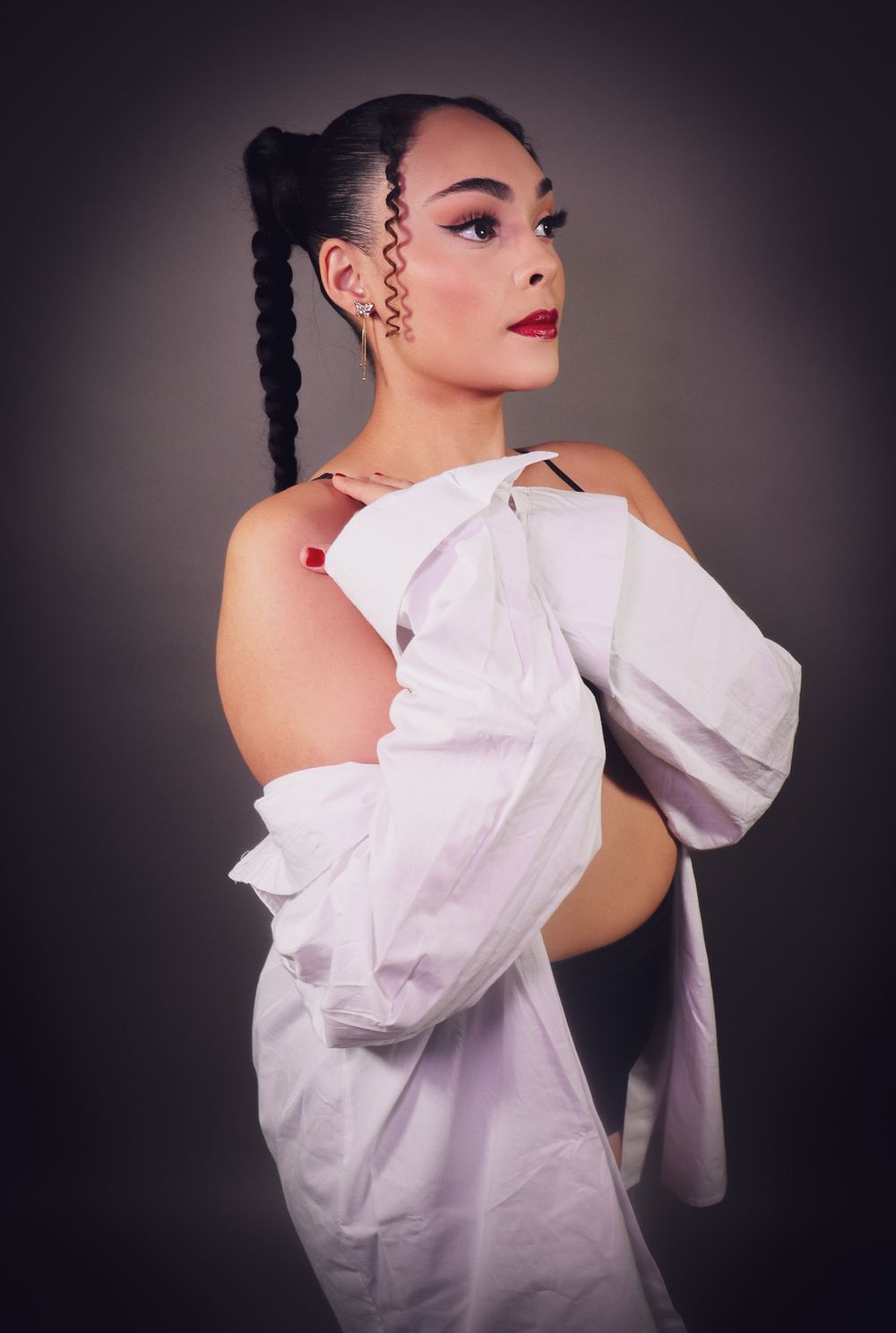 Woman with braided hair and red lips posing in white off shoulder shirt against dark background.