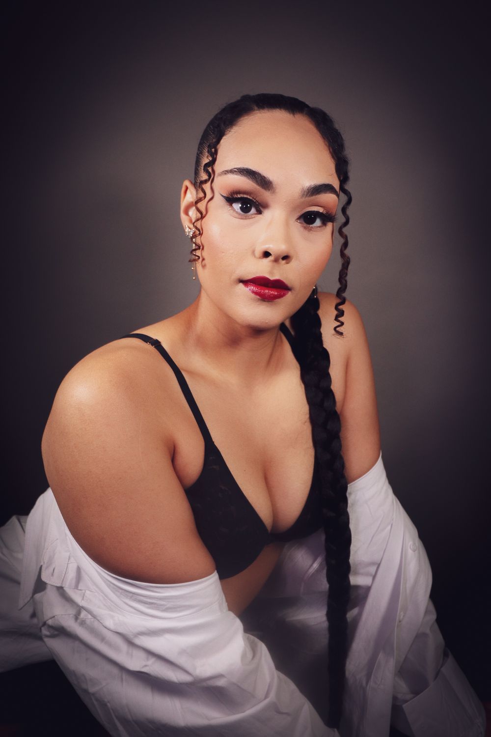 Young woman with long braided hair and red lips poses in black bra and white off-shoulder shirt in studio.