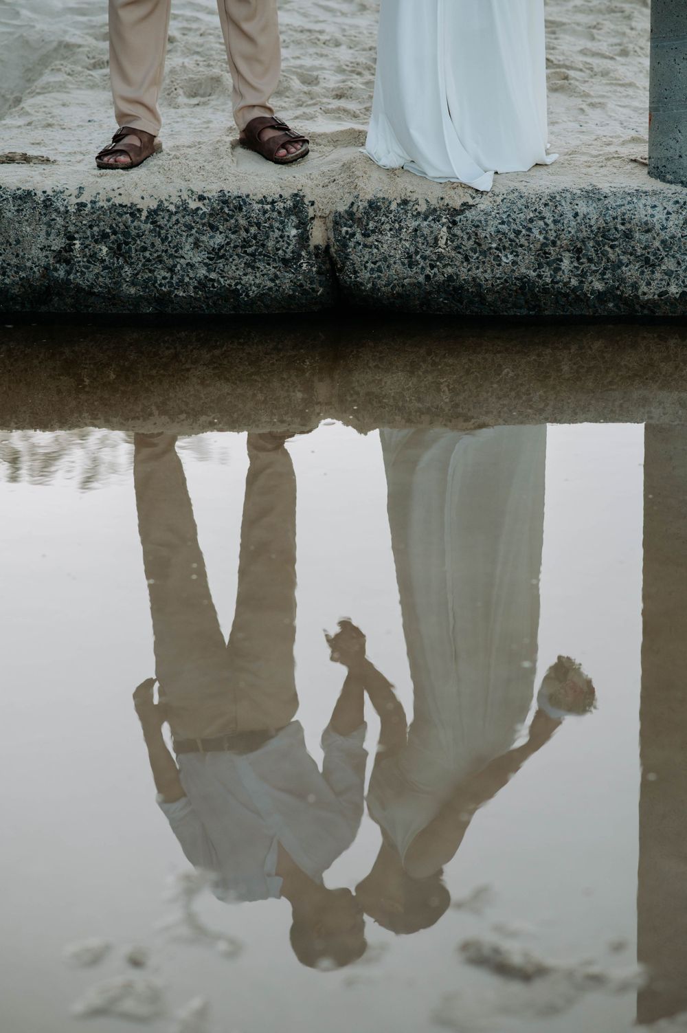 Reflection of a couple's legs and shadows in water while standing on a stone ledge.