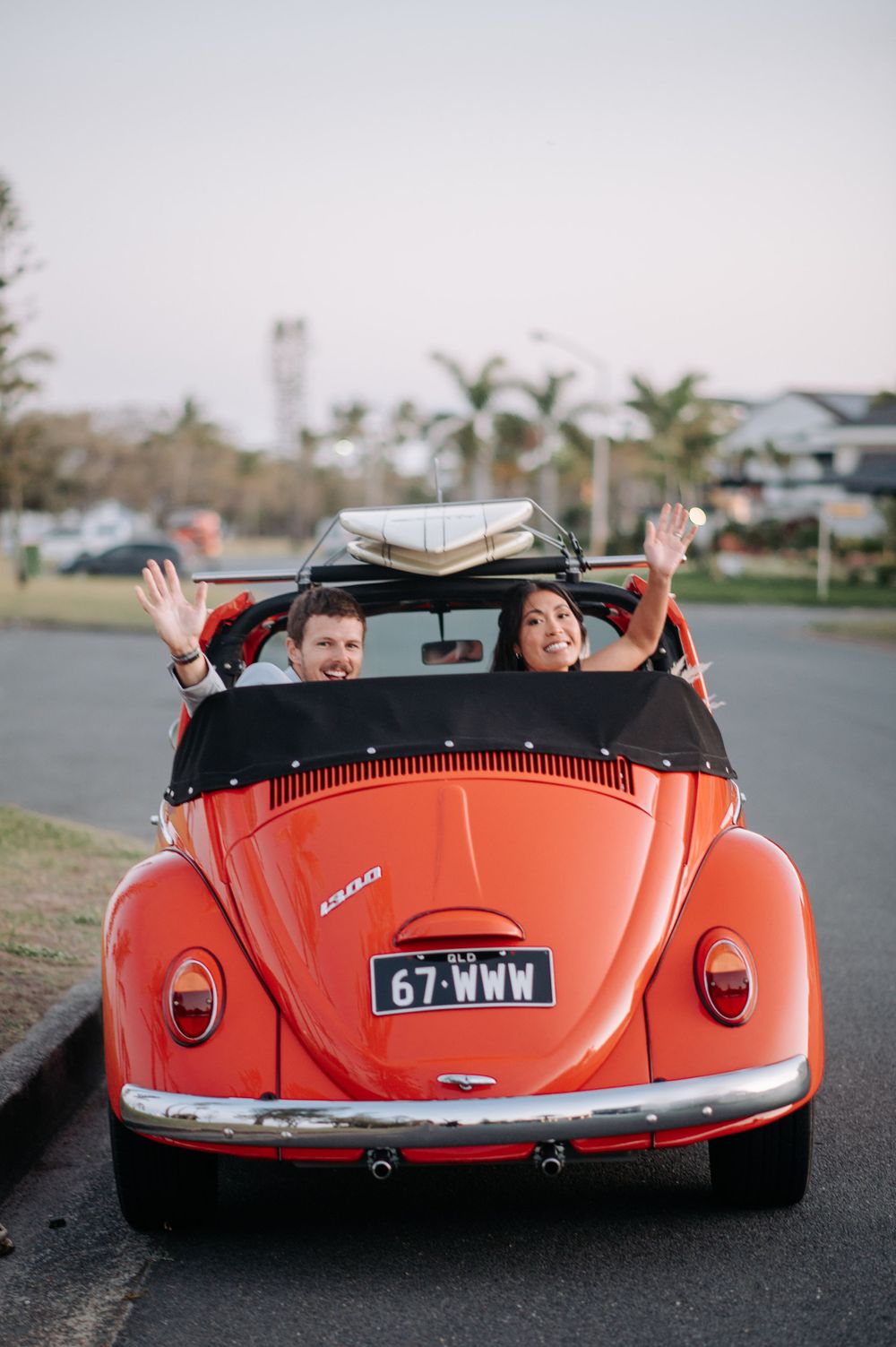 Couple waving from vintage orange Volkswagen Beetle convertible with palm trees in background.