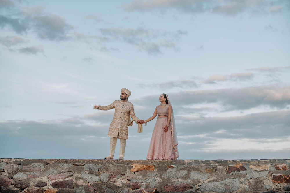 Couple in formal attire holding hands while standing on stone wall against cloudy sky.