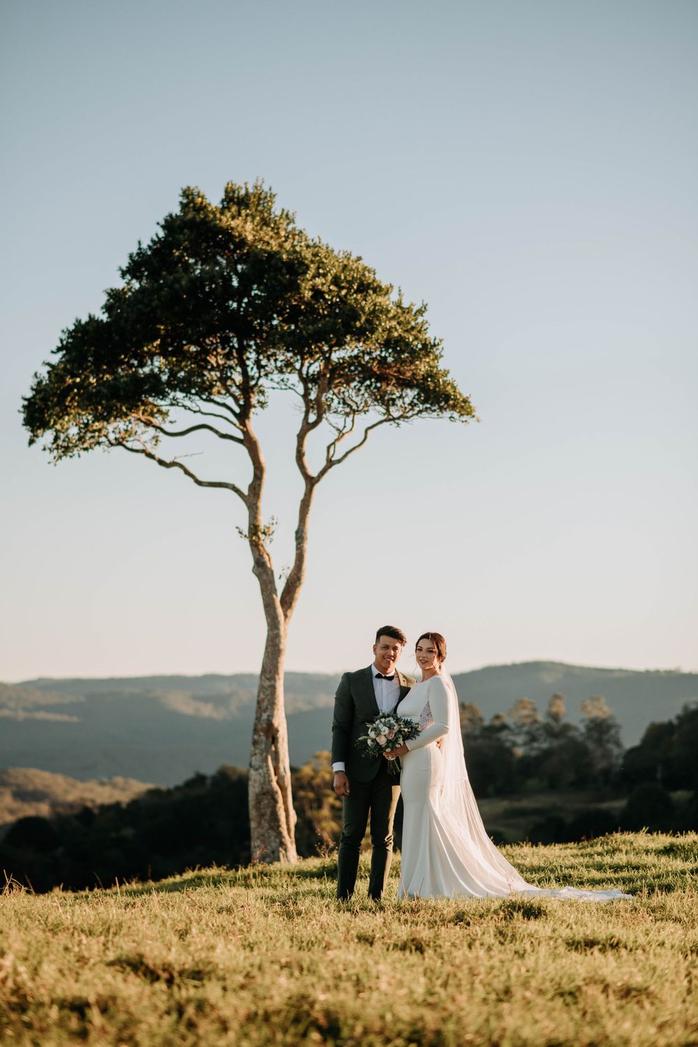 Scenic wedding portrait with couple standing beneath lone tree on hilltop at sunset.