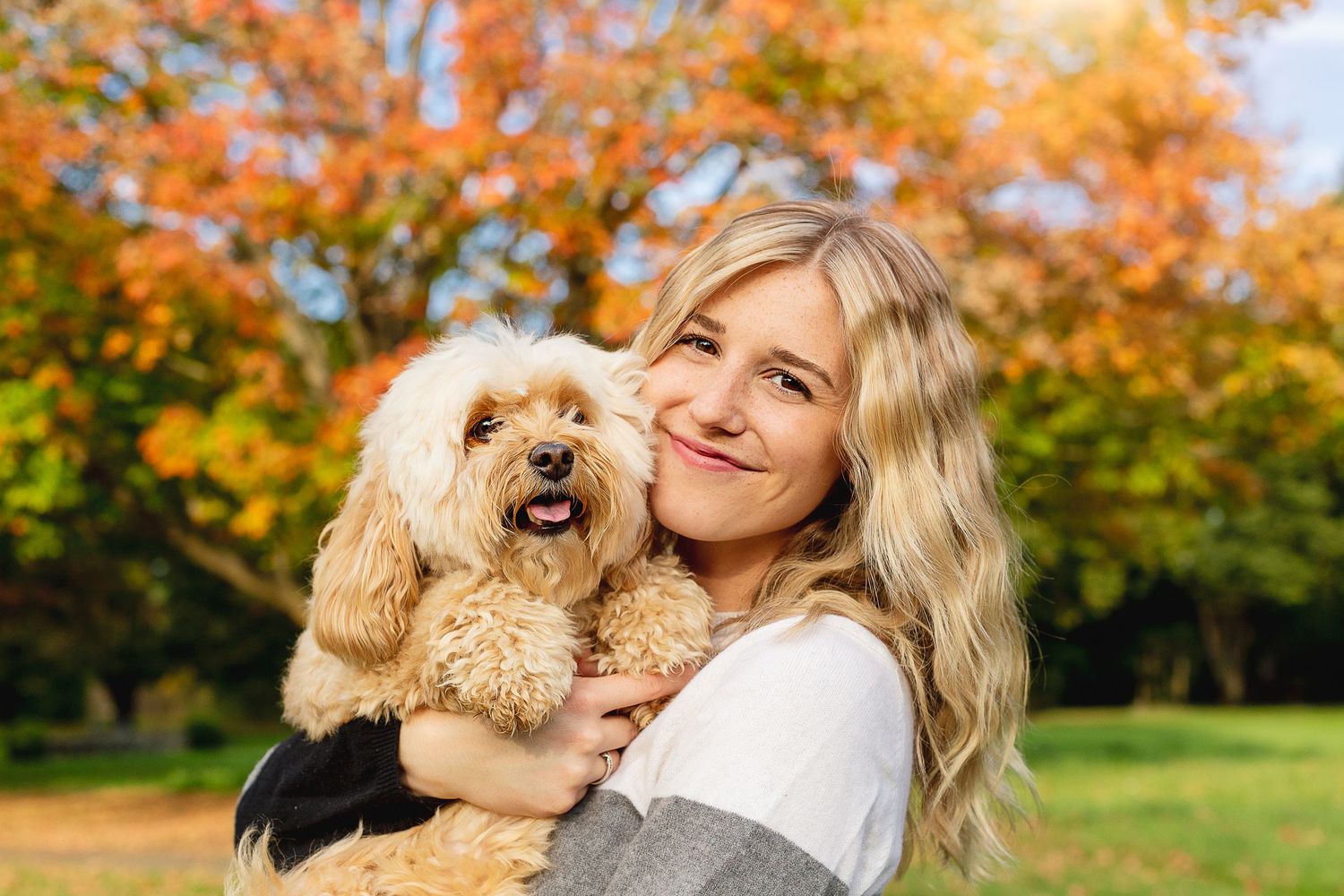 Person holding smiling fluffy dog outdoors with autumn foliage in background.