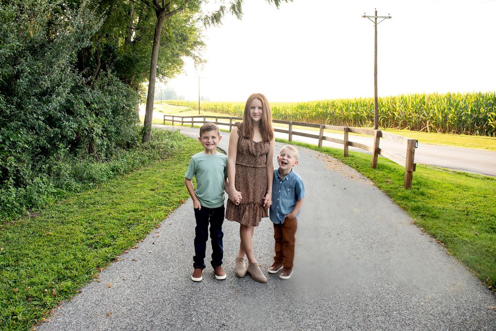 Three people stand together on a rural country road beside cornfields.