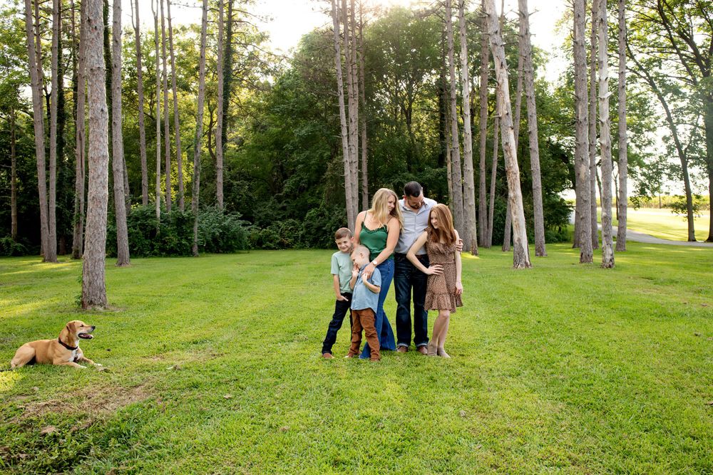 Family stands together with their dog in a grove of tall trees at sunset.