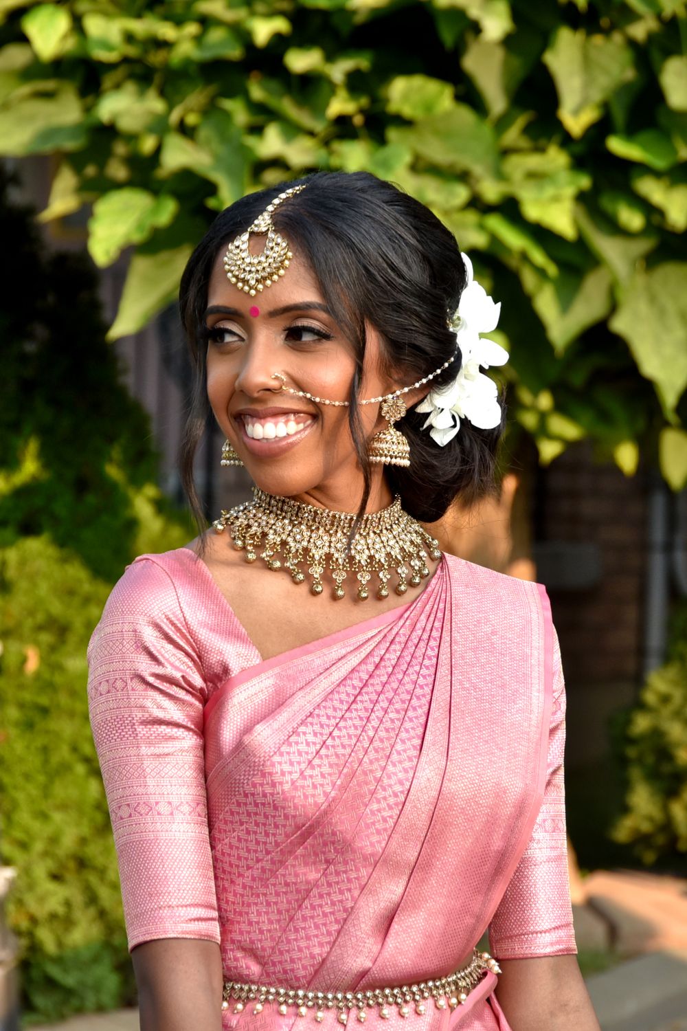 Woman in pink silk saree with traditional Indian bridal jewelry posing outdoors.
