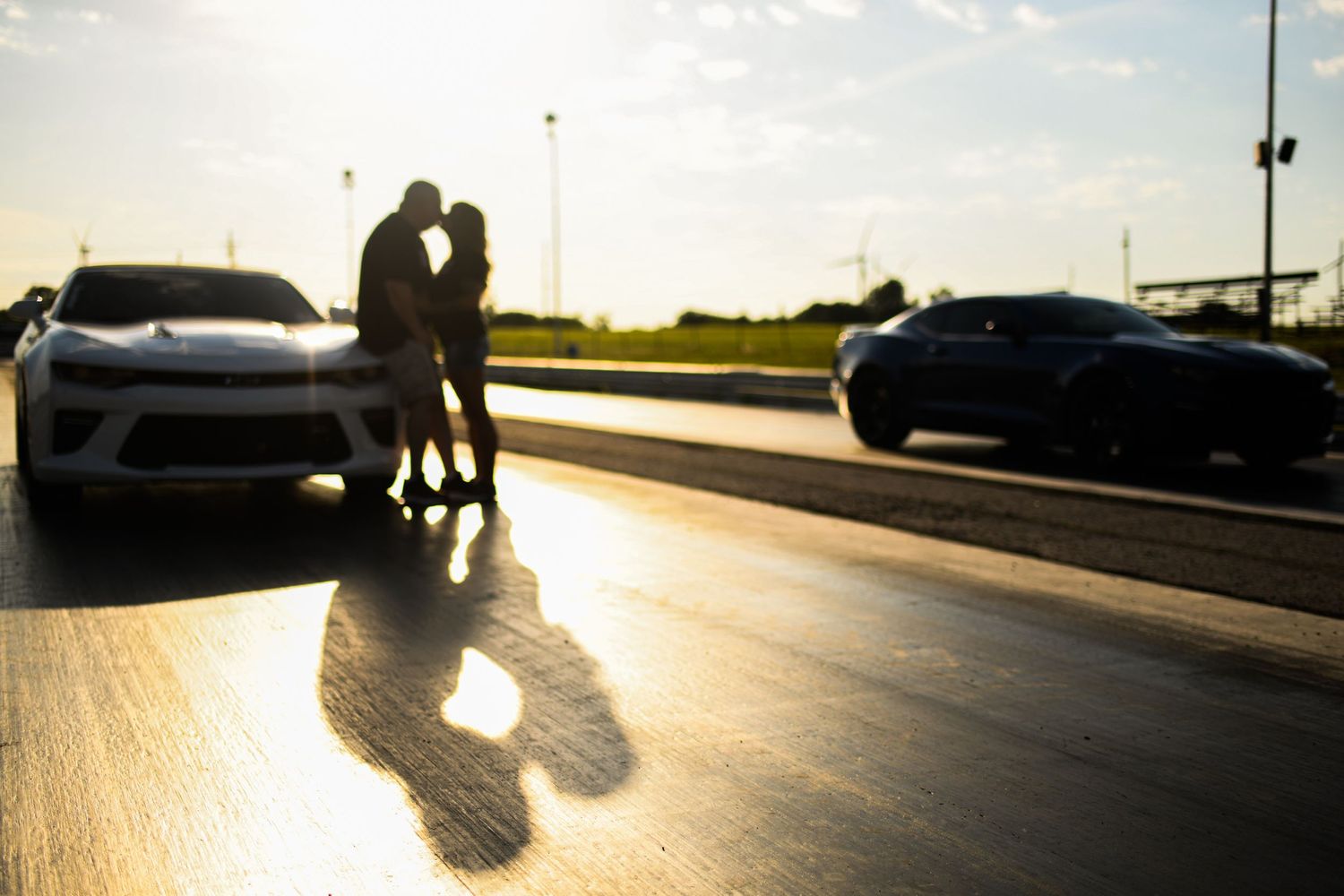 A silhouetted couple stands in golden sunlight between two parked cars on a reflective surface.