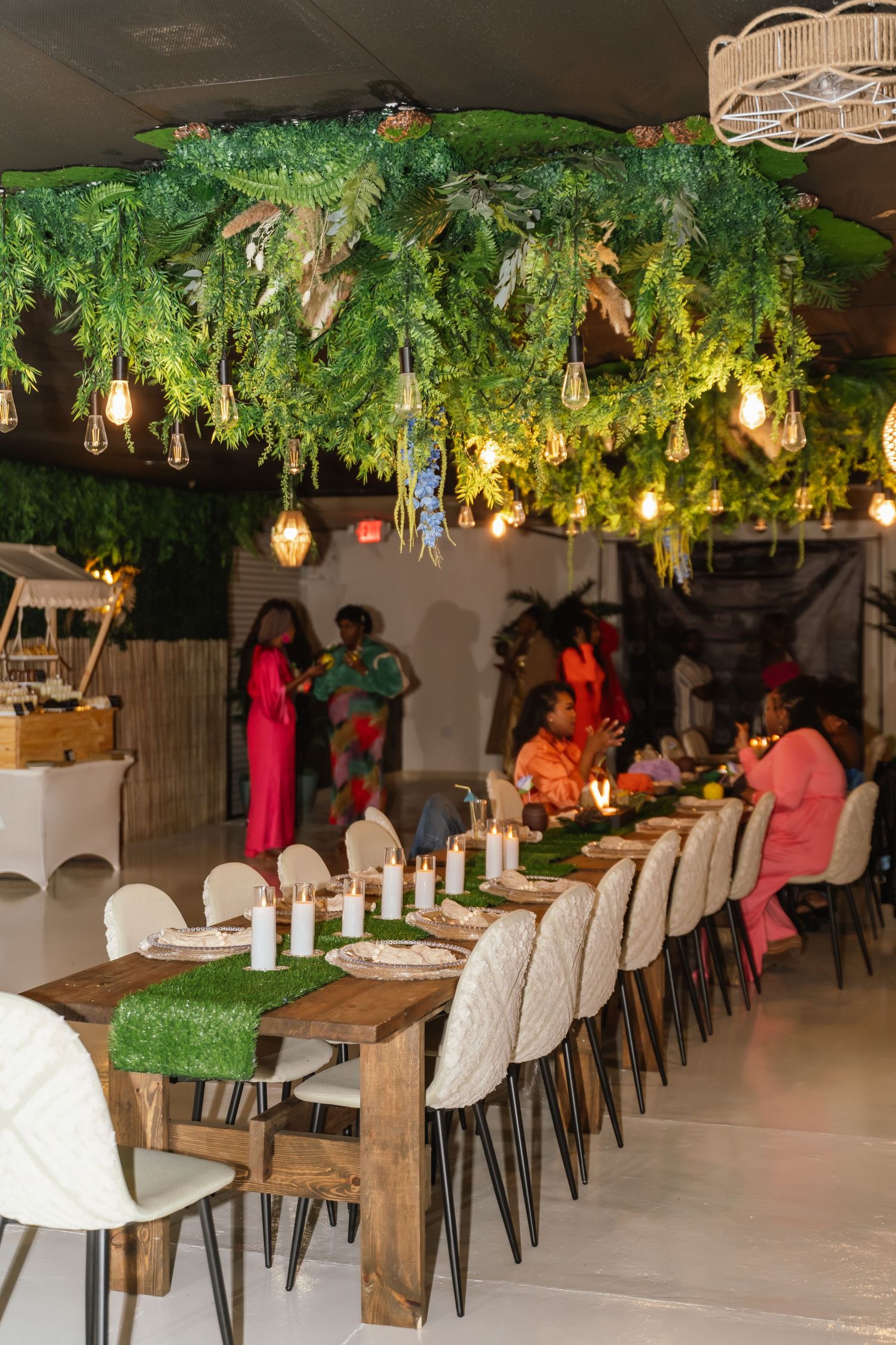 An elegant dining setup with hanging greenery and Edison bulbs over a long wooden table.