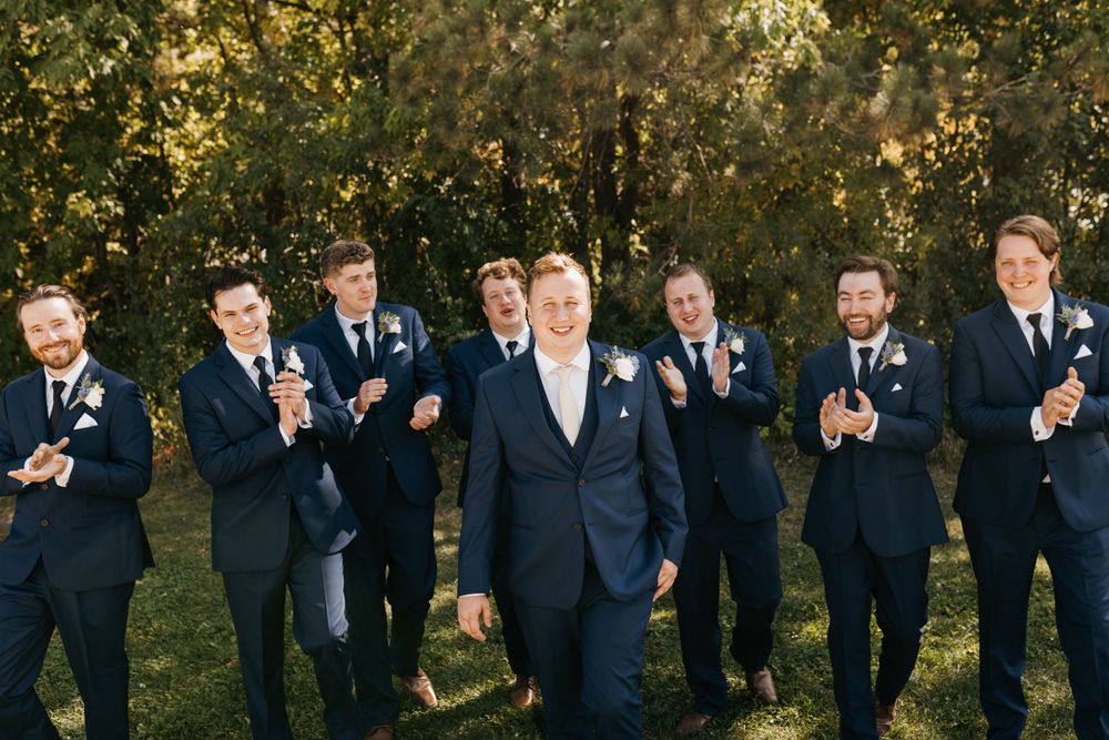 Groom walks confidently as smiling groomsmen in navy suits clap and celebrate outdoors at a wedding.