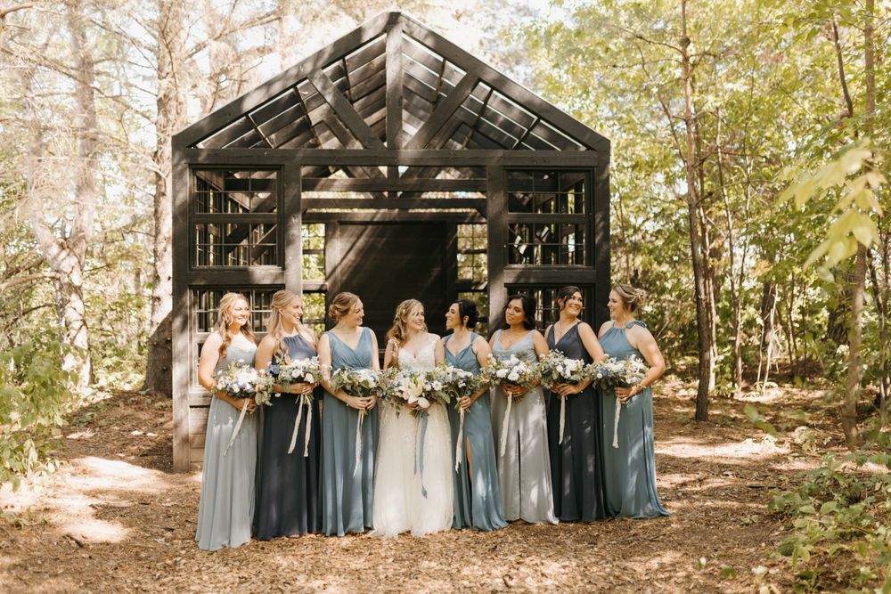 Wedding party of bridesmaids in dusty blue dresses pose outdoors near a dark wooden barn structure.