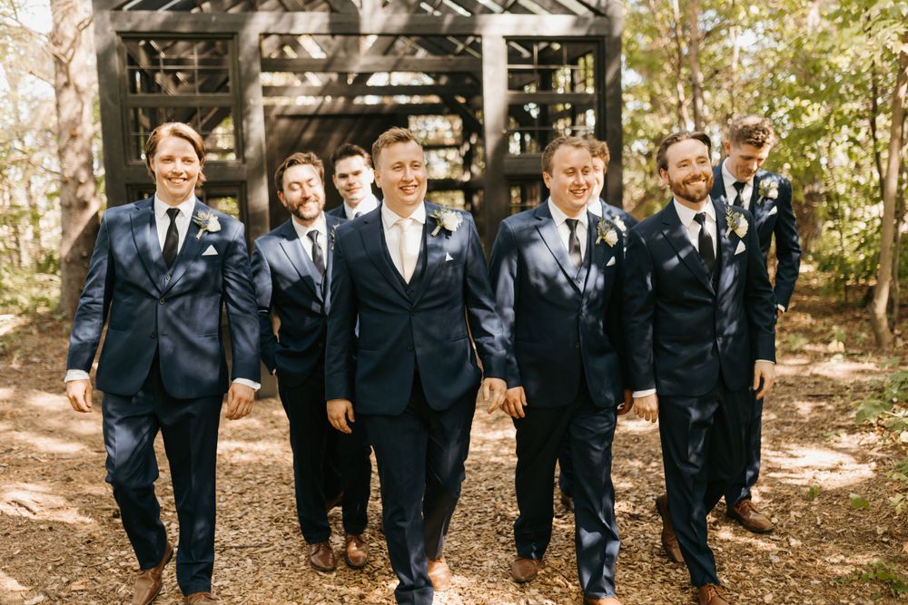 Groom and groomsmen in navy suits walk confidently together through a wooded outdoor wedding venue.