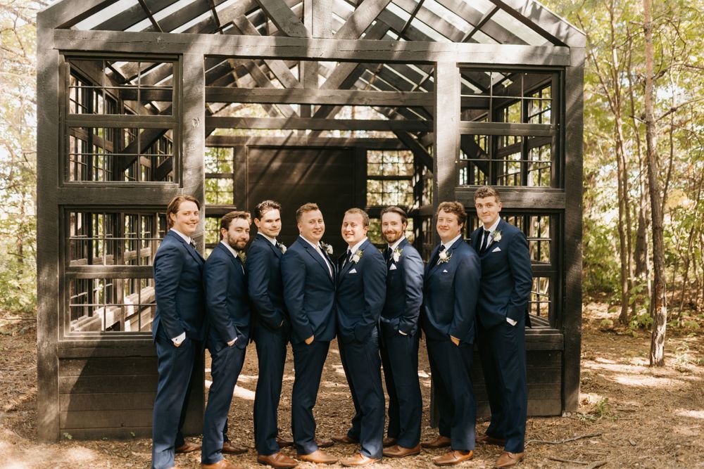 Groomsmen in matching navy suits pose together in front of a rustic wooden greenhouse structure outdoors.