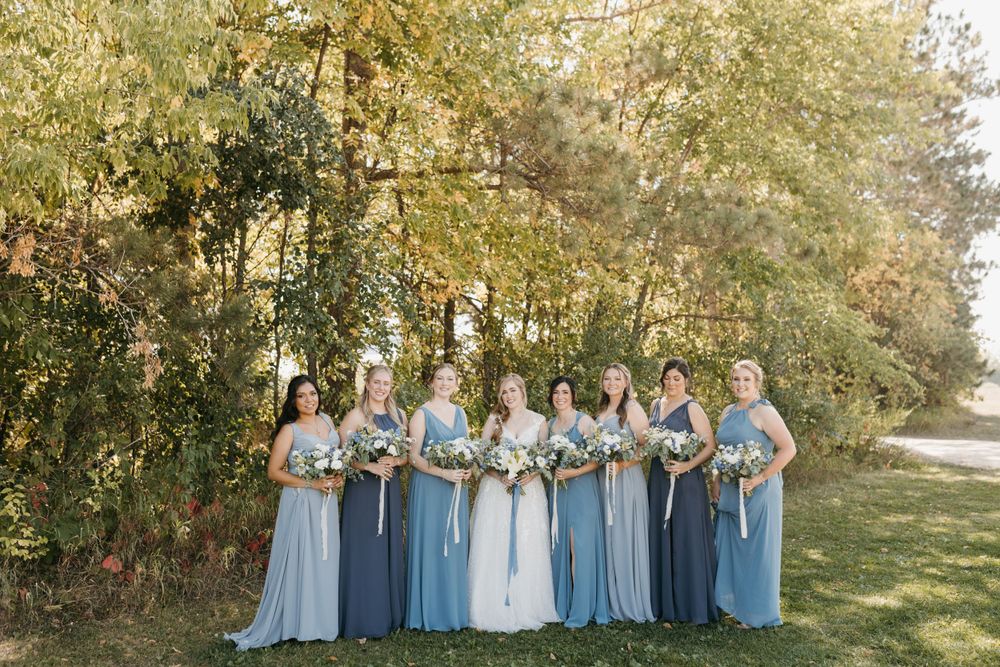 Bride and bridesmaids in blue gowns holding bouquets outdoors among autumn trees.