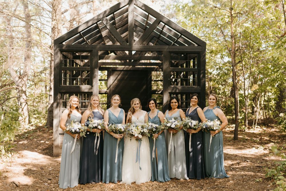 Bride and bridesmaids in mismatched blue gowns pose with bouquets in front of a rustic wooden structure.