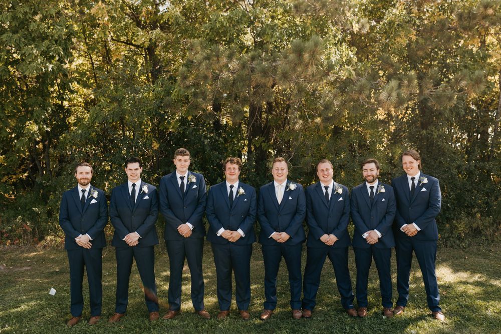 Groom with groomsmen in navy suits posing in front of autumn trees.