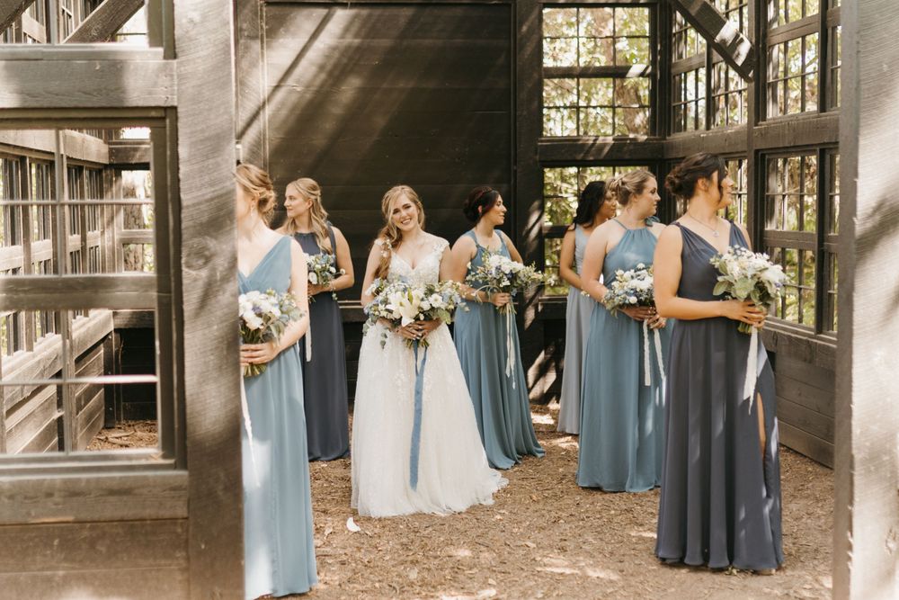 Bride in white gown stands with bridesmaids in blue dresses inside a rustic wooden barn venue.