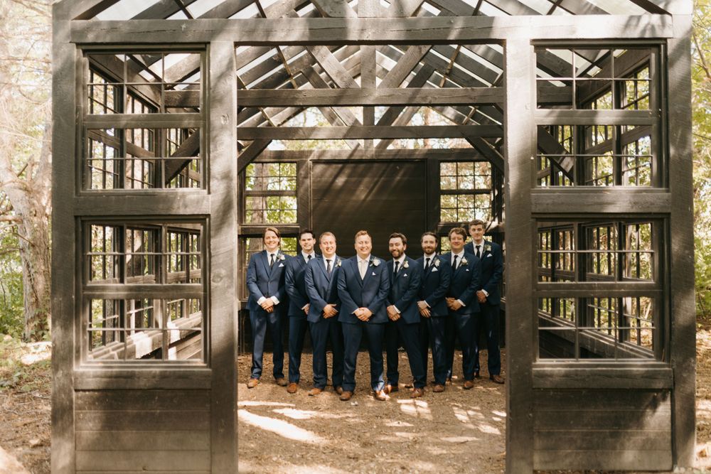 Groom and groomsmen in navy suits pose inside a rustic wooden greenhouse structure at a wedding venue.