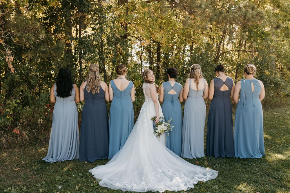 Bride and bridesmaids showing back of blue dresses in front of autumn trees.