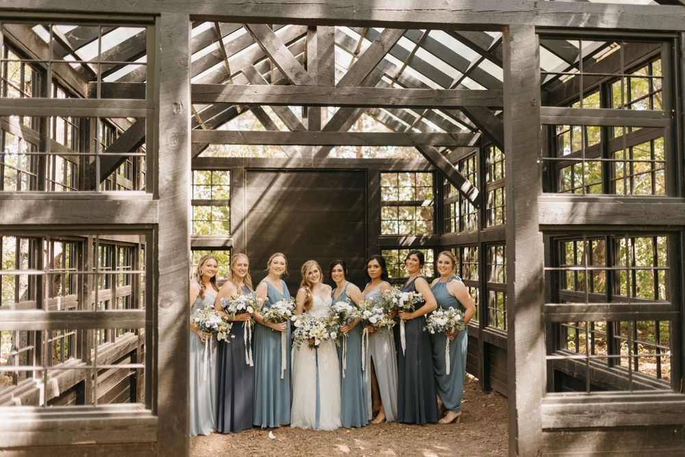 Bride and bridesmaids in shades of blue pose inside a rustic wooden greenhouse structure in the woods.