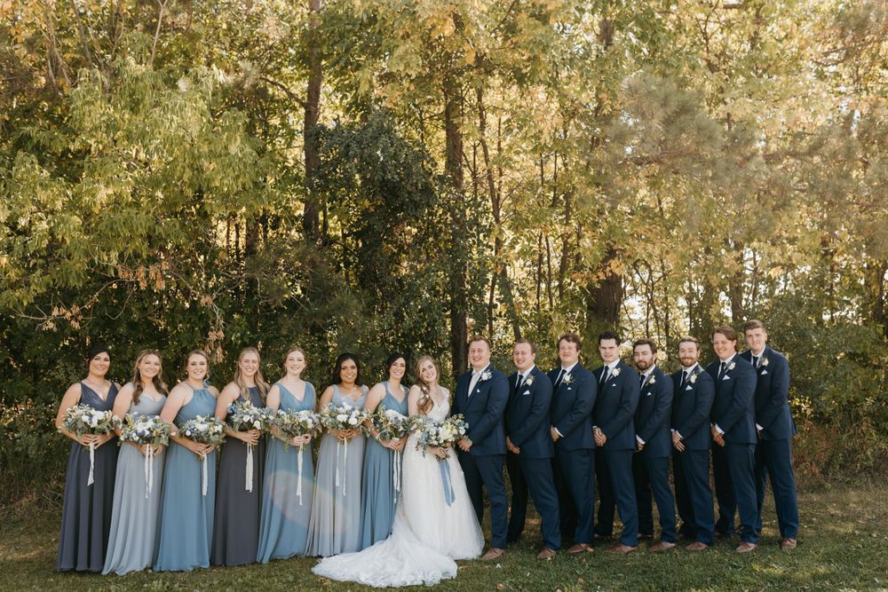 Full wedding party with groomsmen in navy suits and bridesmaids in blue gowns outdoors.