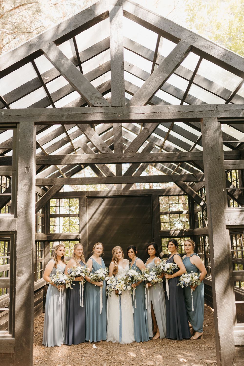 Bridesmaids in mismatched blue gowns hold bouquets while posing under a rustic wooden pergola structure.