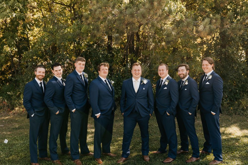 Groom and groomsmen smiling in navy suits with boutonnieres among fall foliage.