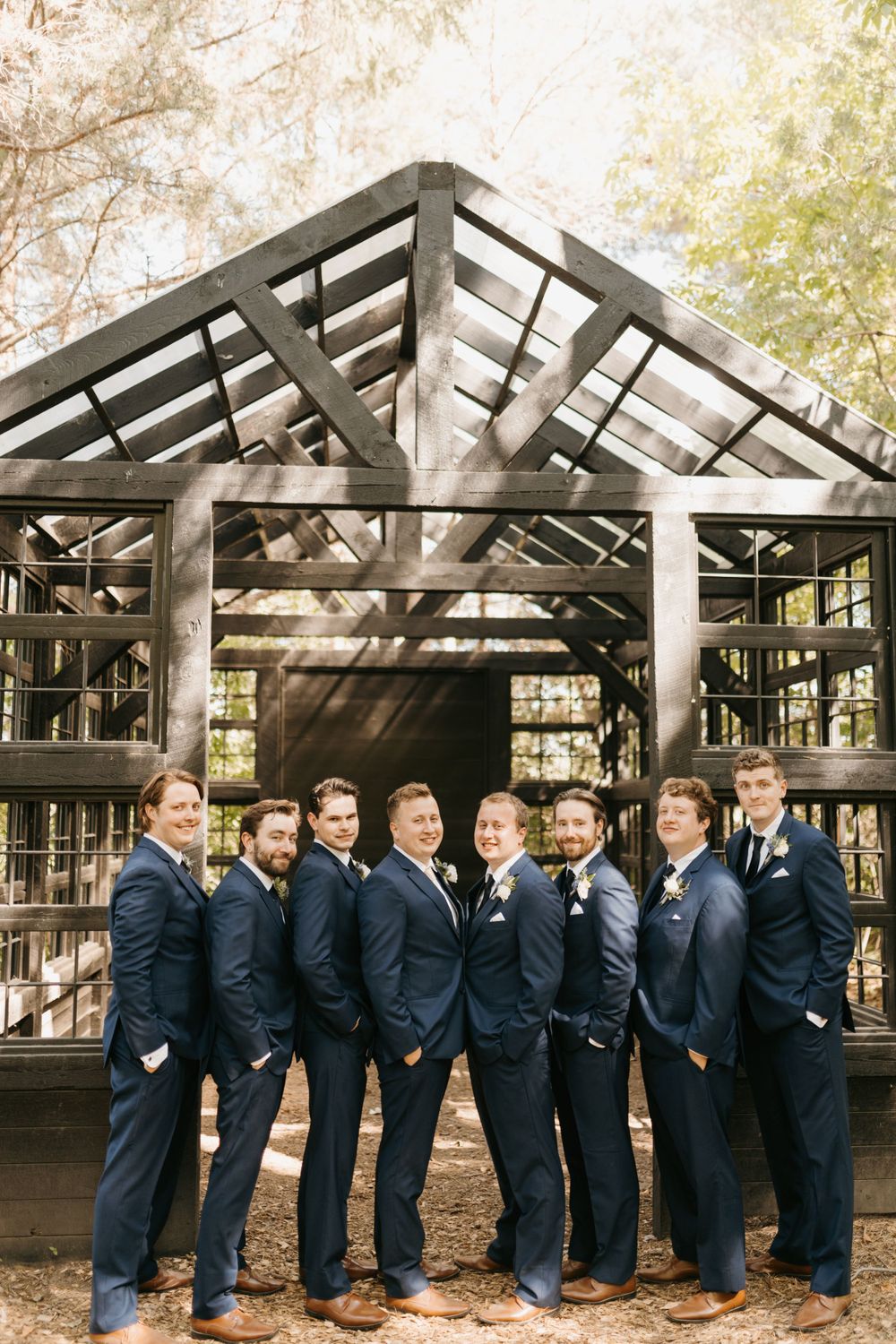 Groomsmen in navy suits and brown shoes pose in front of a rustic wooden pavilion in a wooded setting.