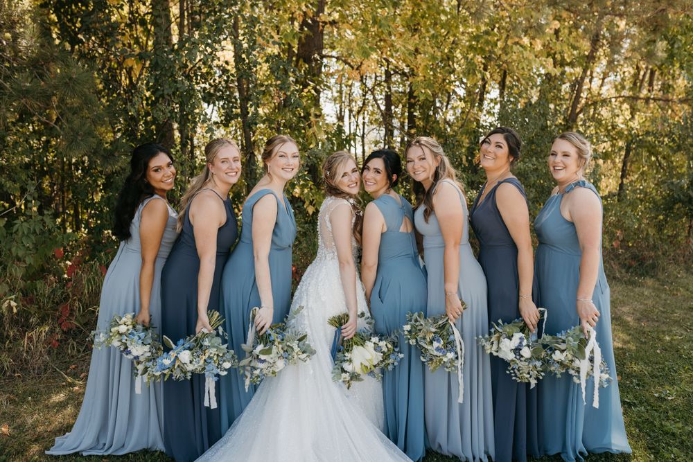Bride in white gown with bridesmaids in dusty blue dresses laughing outdoors.