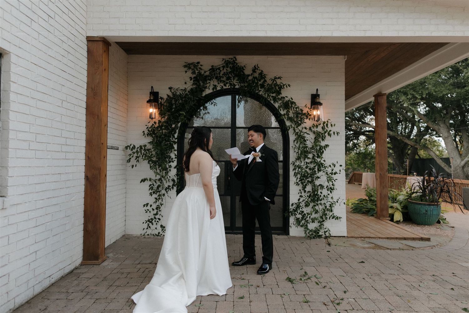 Wedding couple stands beneath a floral arch at a white brick chapel entrance.