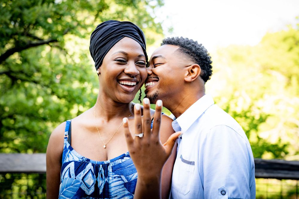 Joyful Black couple laughing together outdoors, woman showing engagement ring while man whispers in her ear.