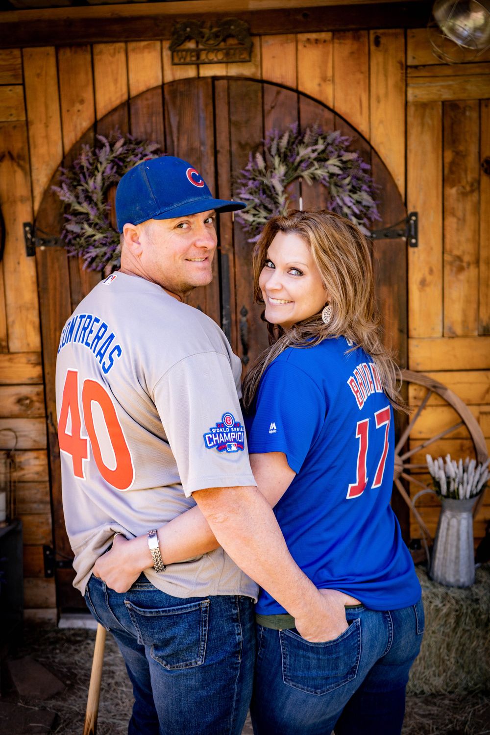 Couple wearing Chicago Cubs baseball jerseys pose together in front of rustic wooden barn doors.
