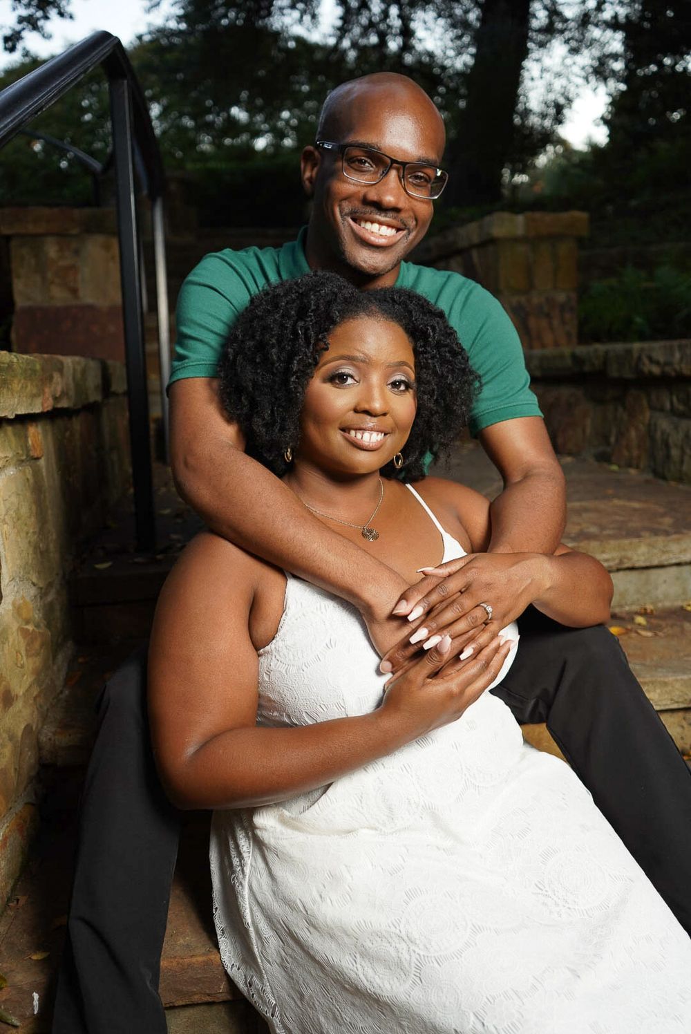 Happy Black couple posing on stone steps, man in green shirt embracing woman in white wedding dress.