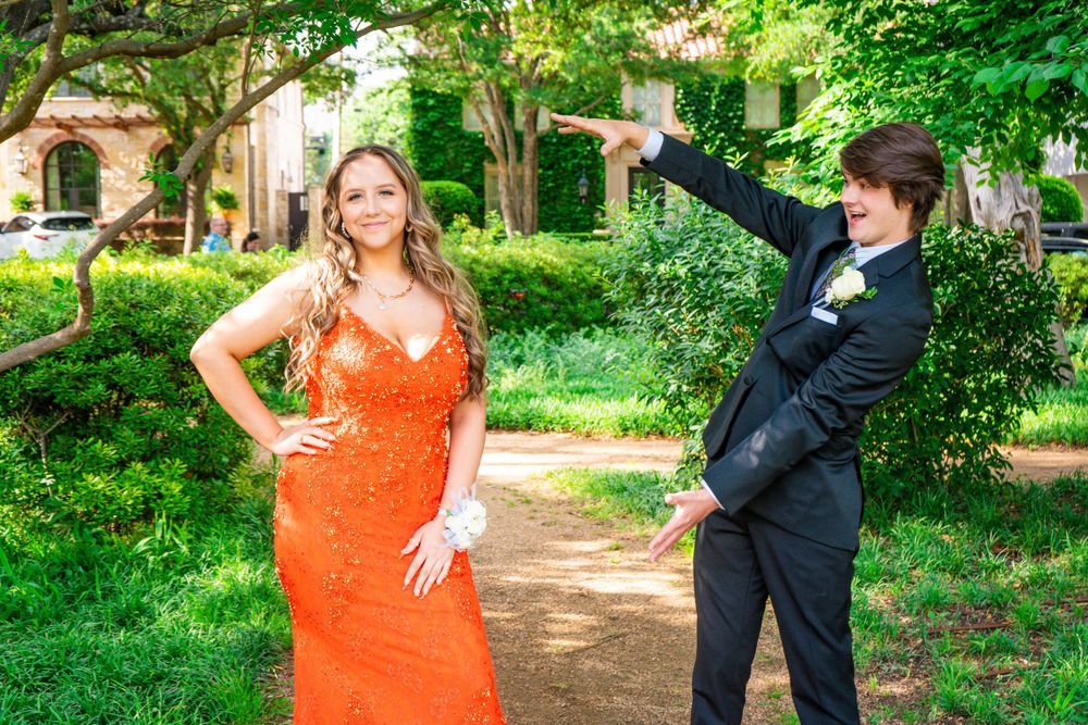 Teen couple in prom attire pose playfully outdoors, girl in orange sequin gown with corsage.