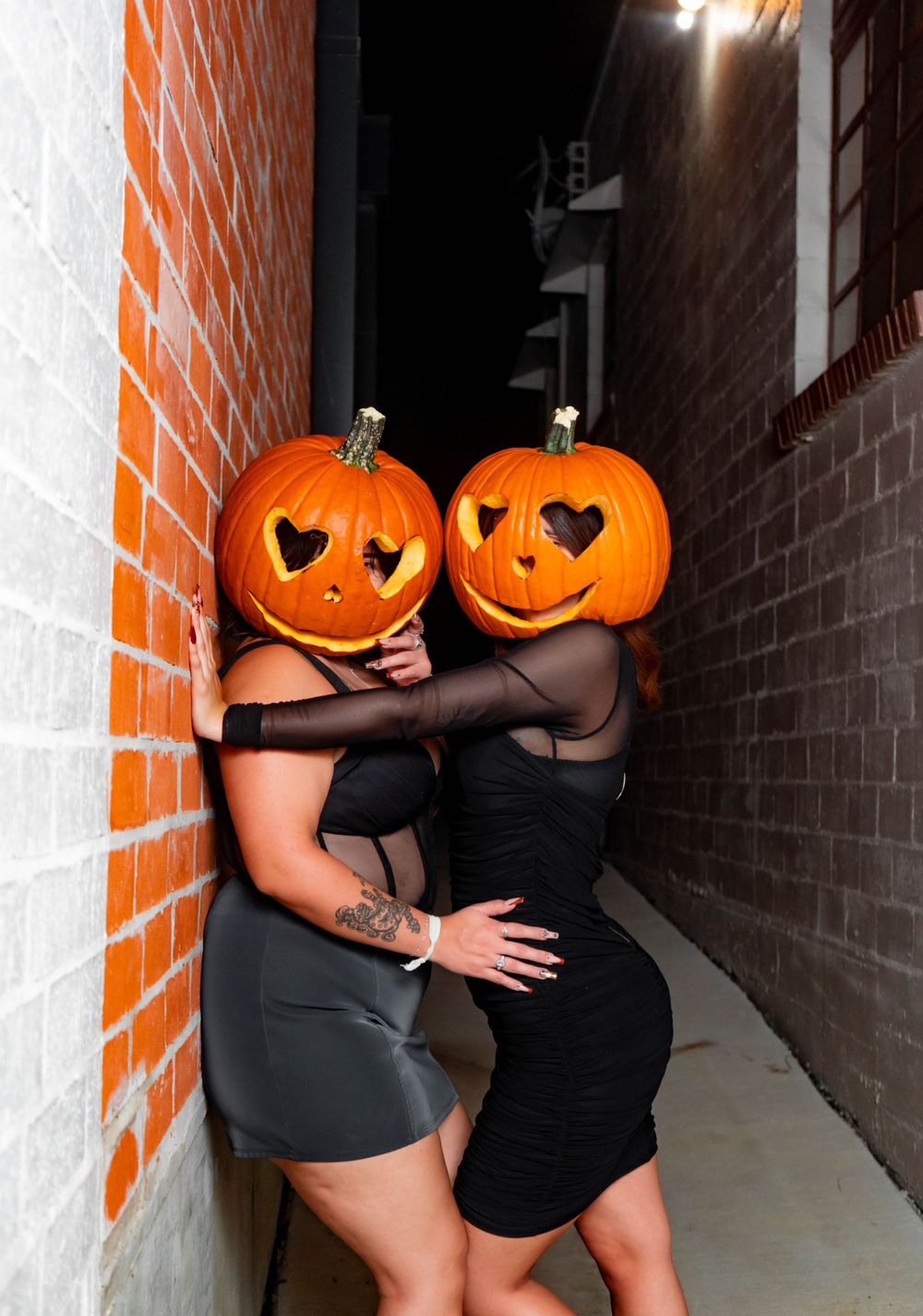 Two women wearing Halloween pumpkin heads pose together in a narrow brick alleyway at night.