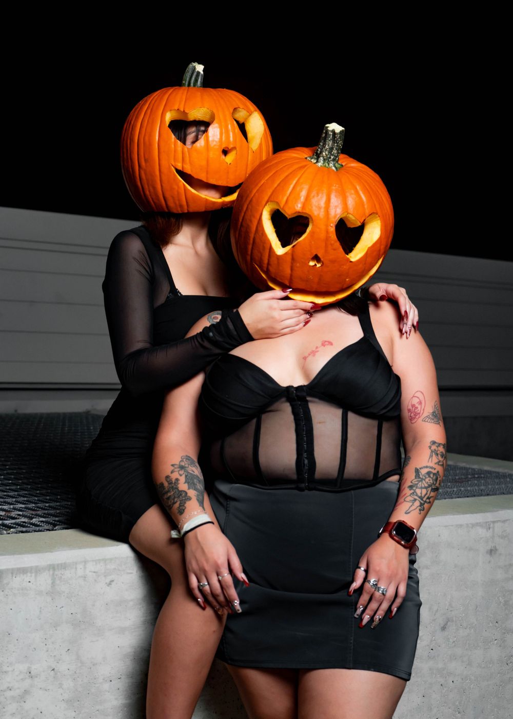 Two tattooed women in black dresses wearing carved pumpkins on their heads embrace outdoors at night.