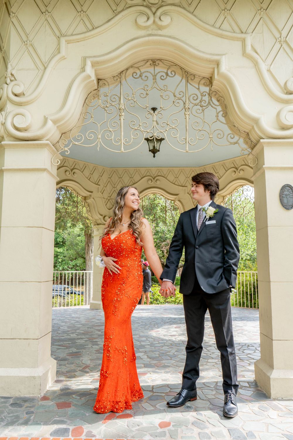 Young couple in formal attire pose under ornate arch, woman in orange sequin gown for prom photos.