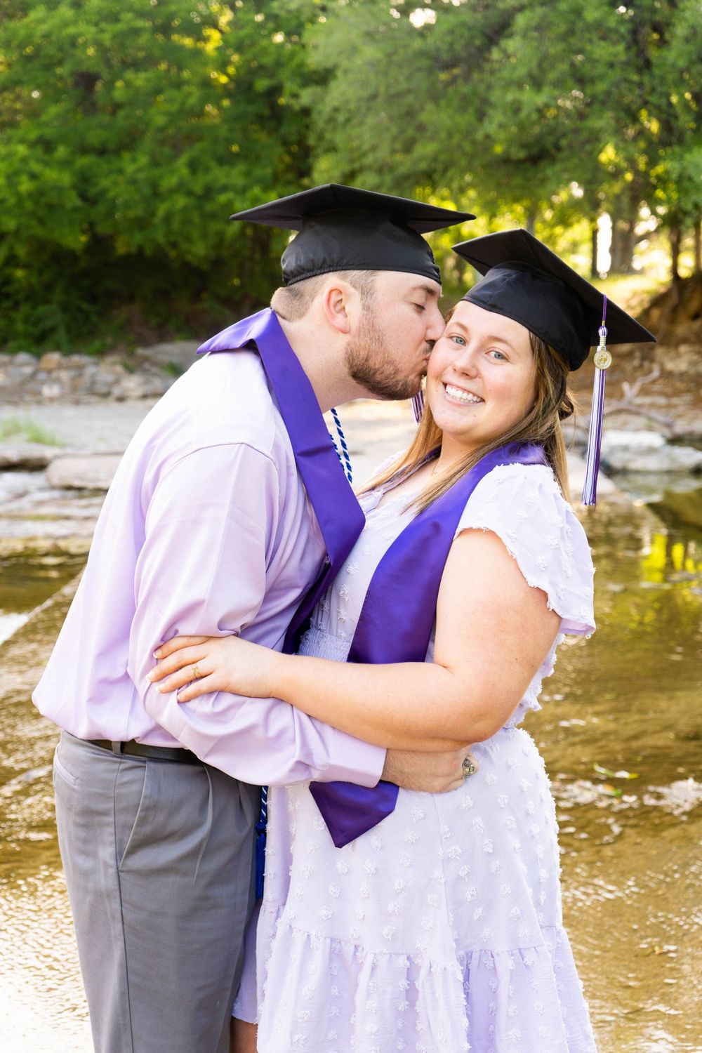 Graduating couple in caps and purple stoles share a kiss near a creek in outdoor photo session.