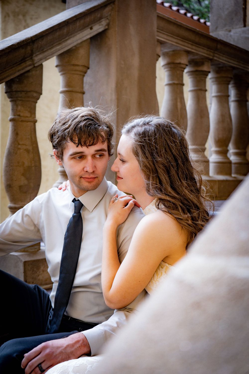 Romantic couple sitting on stone steps, man in white shirt and tie gazes at woman in wedding dress touching his shoulder.
