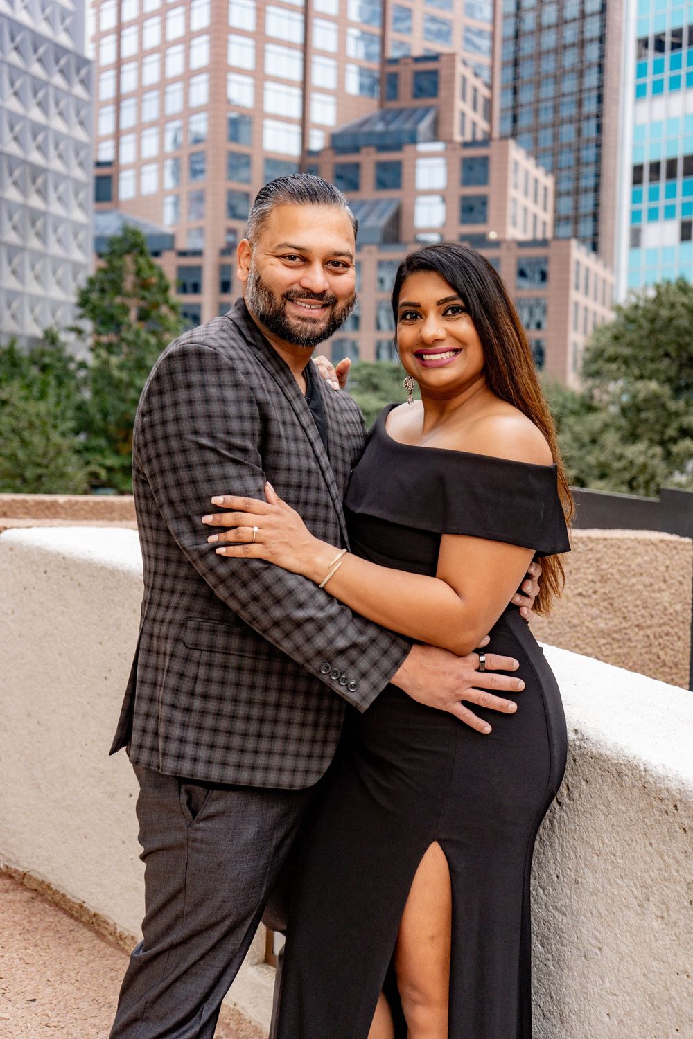 Smiling couple in black and plaid outfits embracing with city skyline in background.