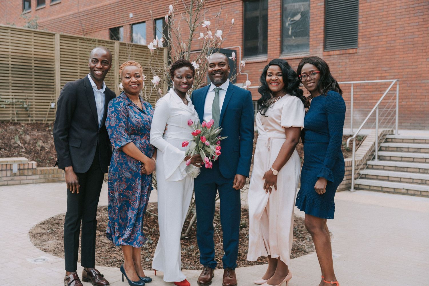 A group in formal attire poses together outside a brick building on a sunny day.
