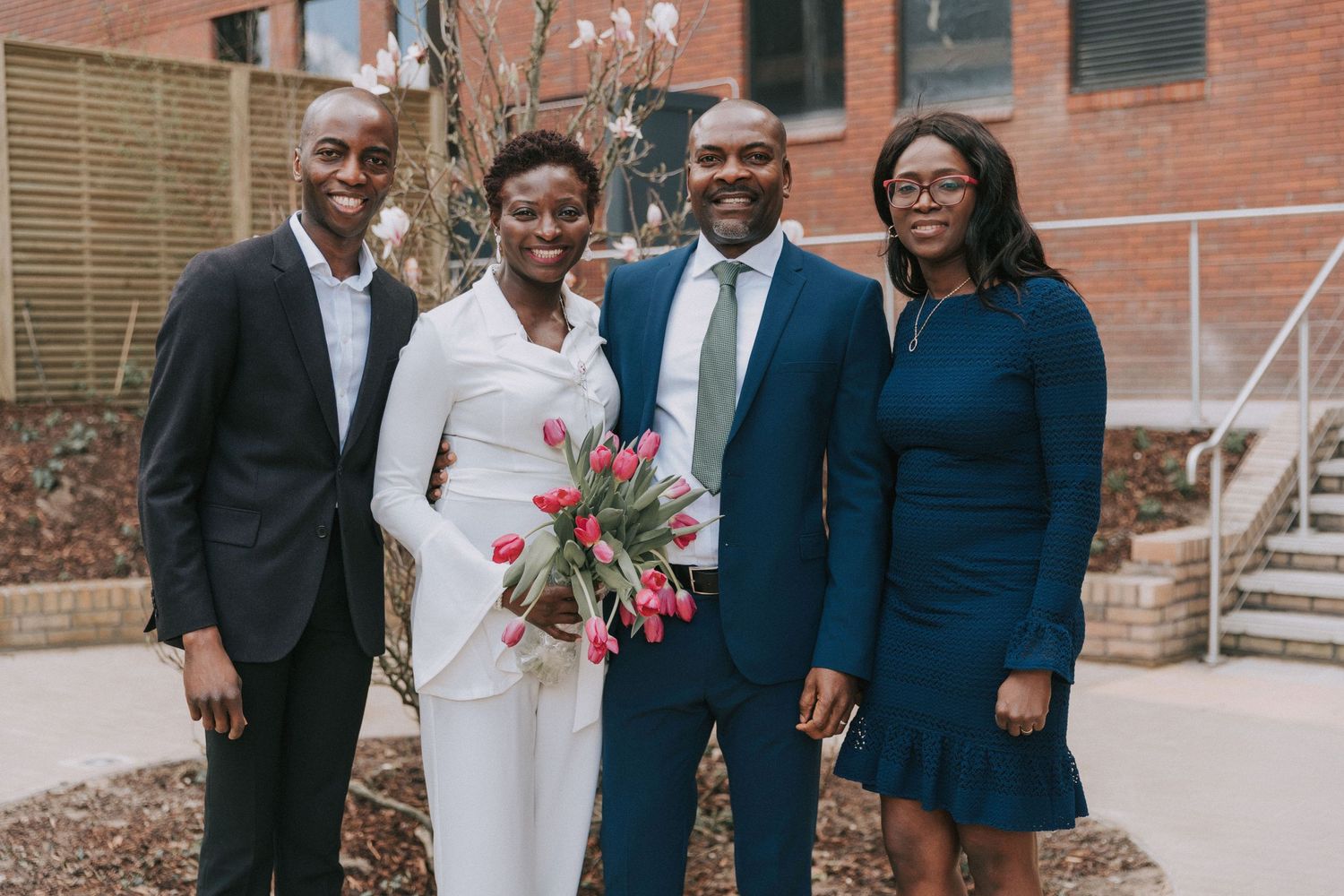 A celebration gathering takes place outside a brick building with multiple people in formal wear.