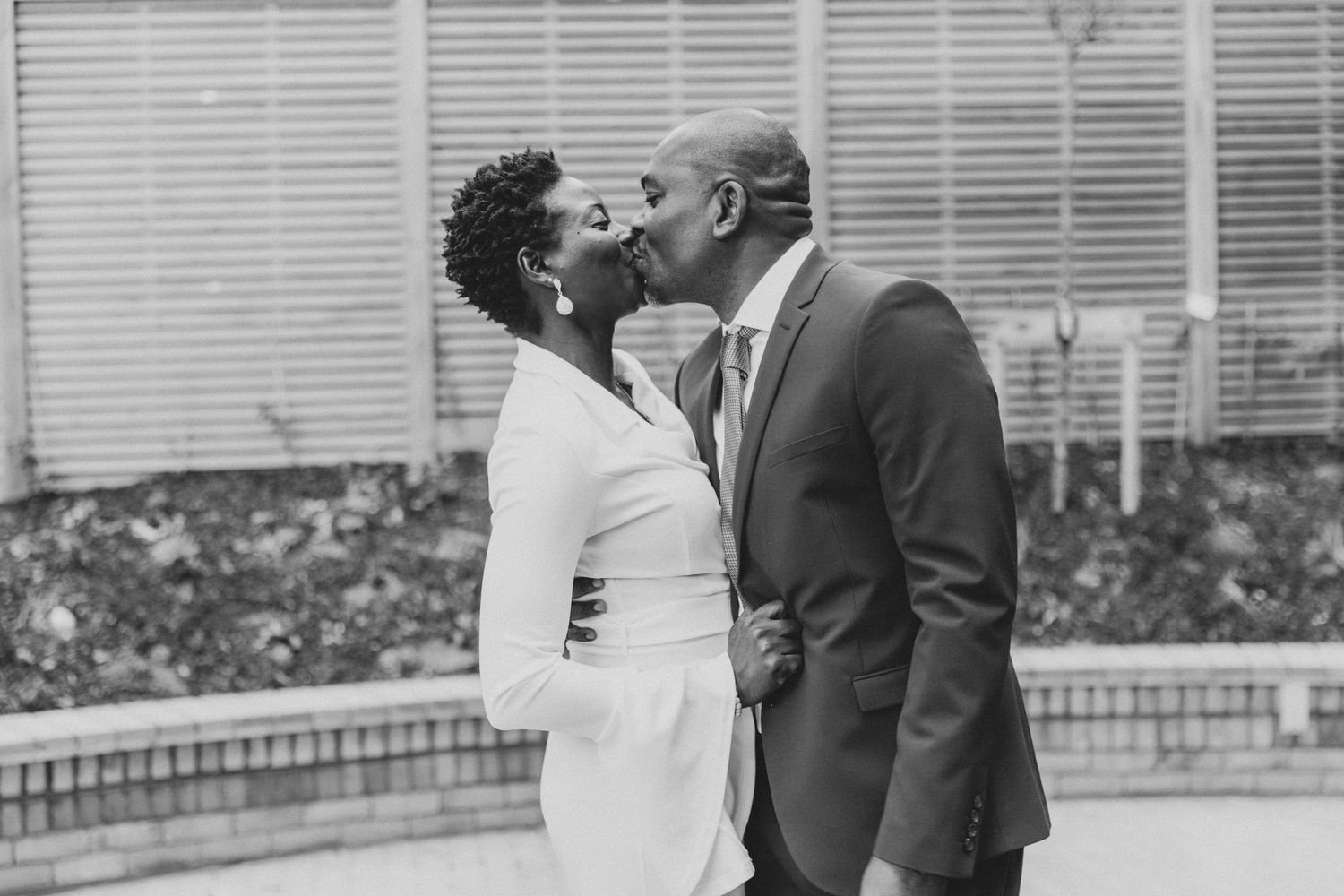A couple in formal attire stands together near a building with white blinds in the background.
