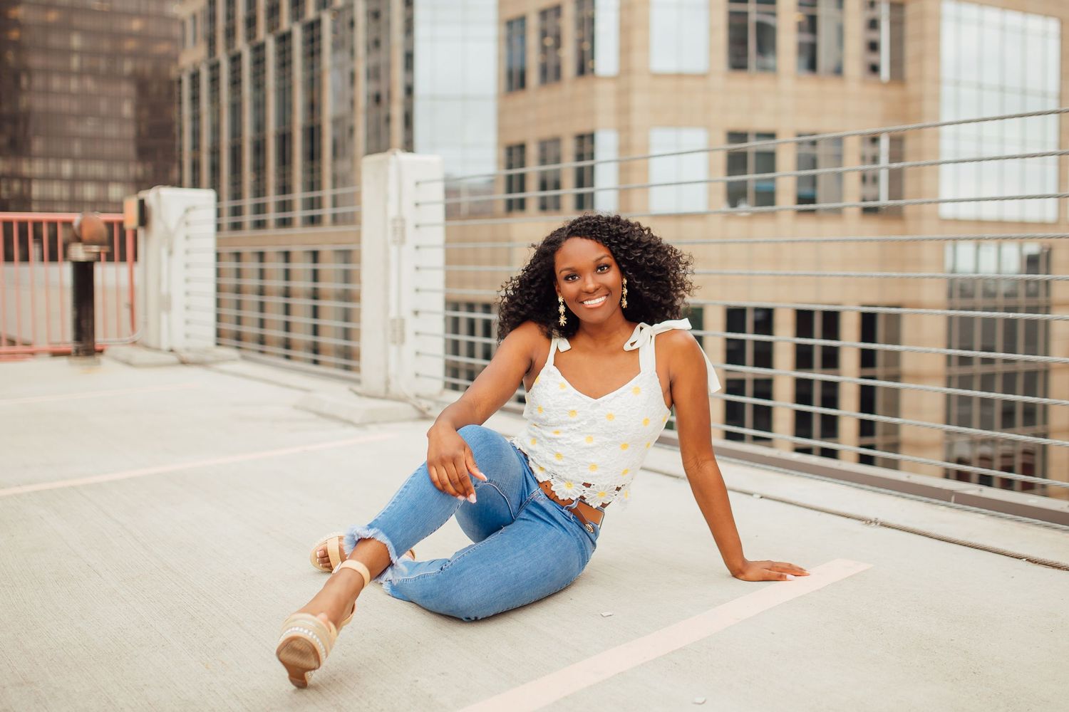 Woman in casual denim sits confidently on an urban rooftop with city buildings in background.
