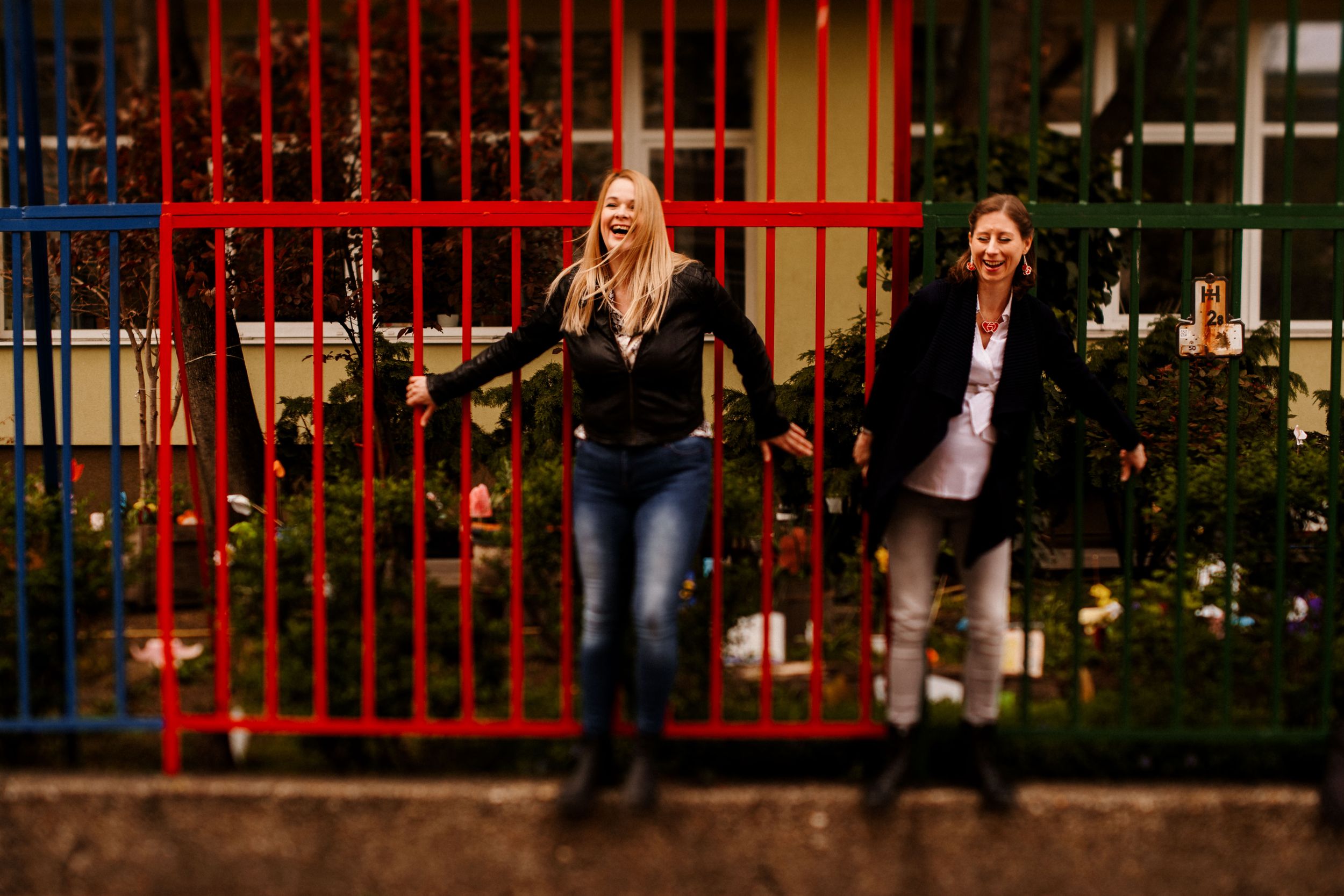 Two people running playfully along a red metal fence at dusk in an urban setting.