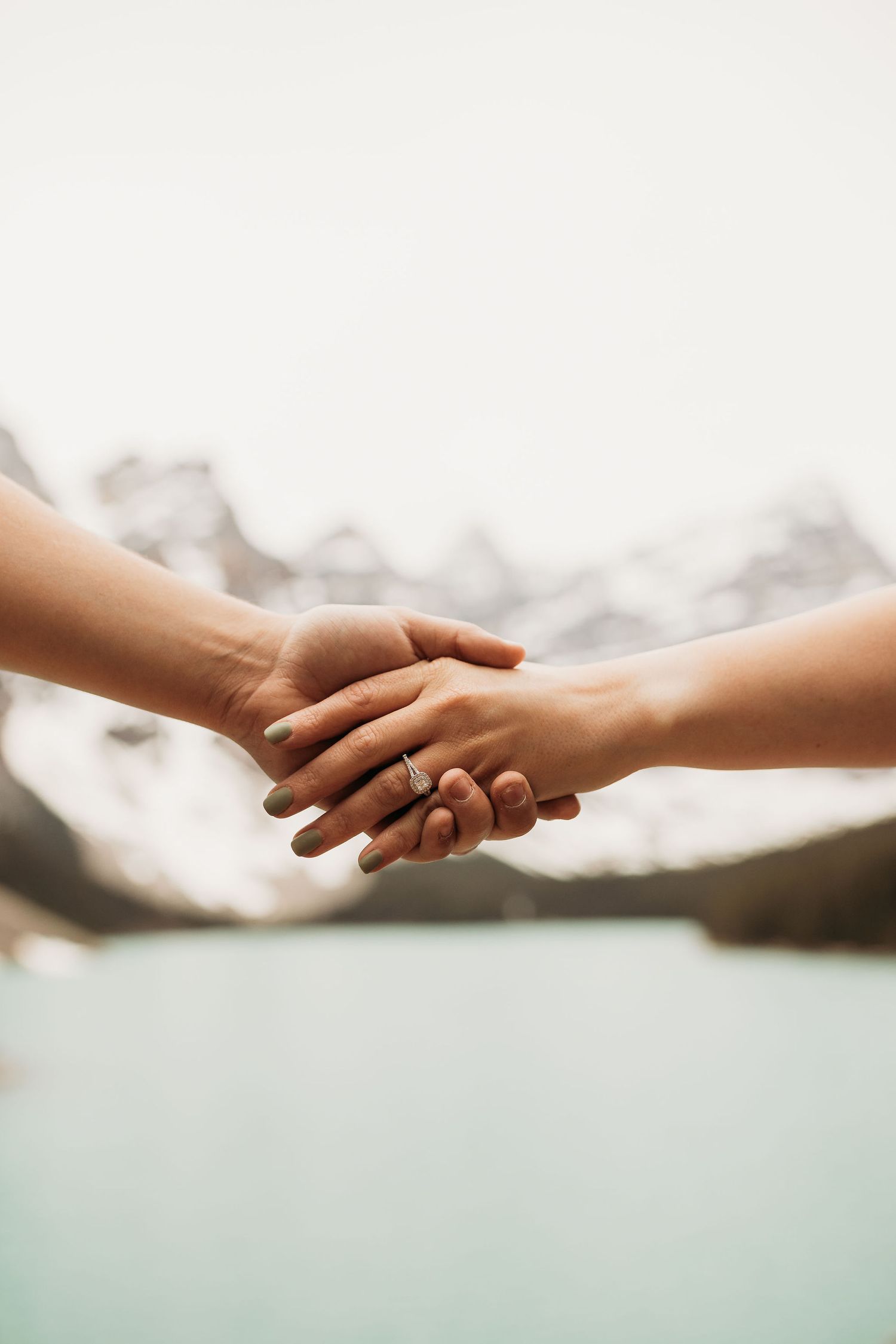 Close-up of hands with engagement ring against mountain lake backdrop.