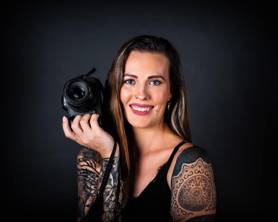 Portrait of Shelby Calahan Leathers,  Caucasian woman in her 30ies, with brown hair and tattoos, smiling with camera.