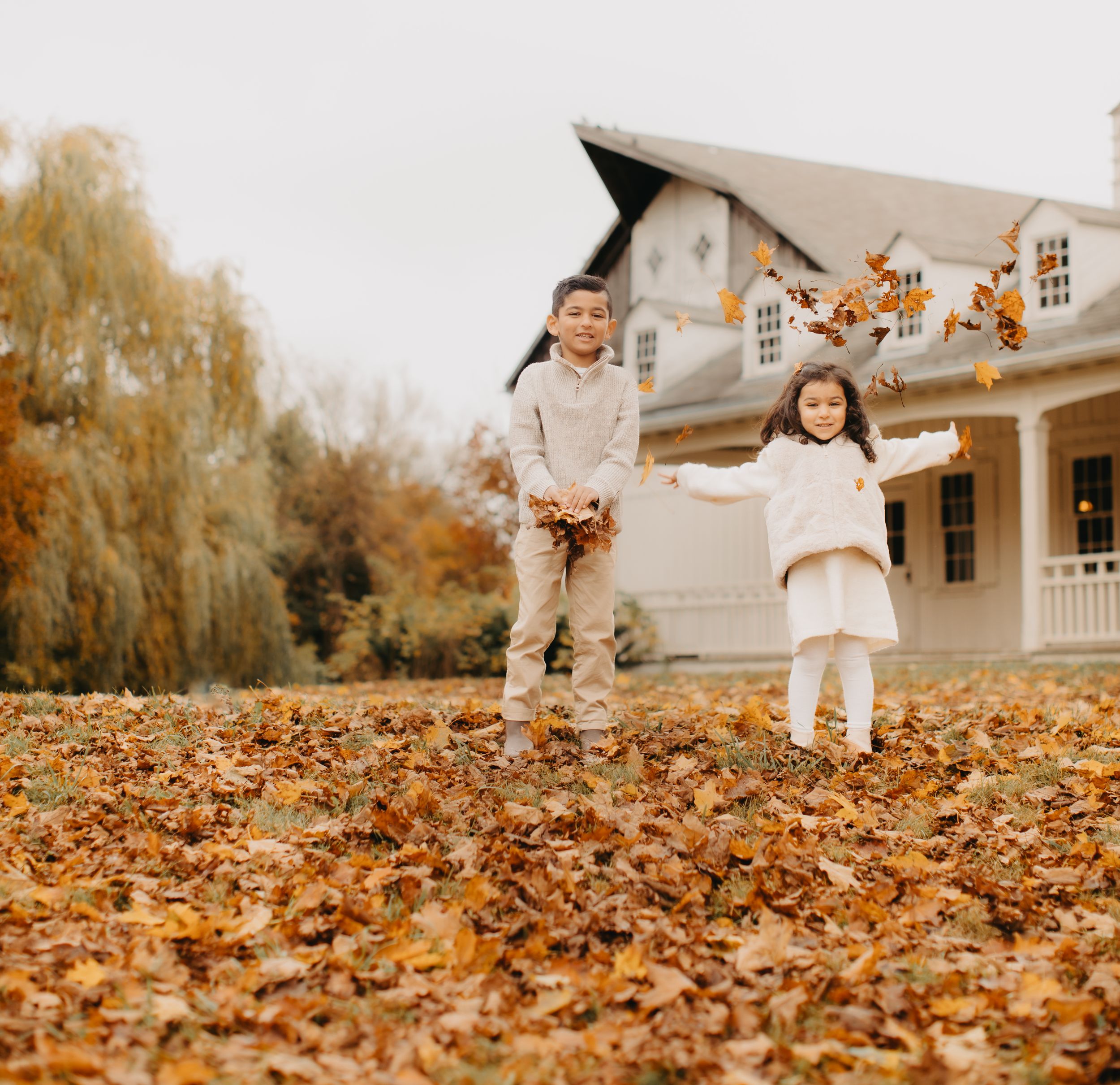 Children playing in fallen autumn leaves near white colonial house.