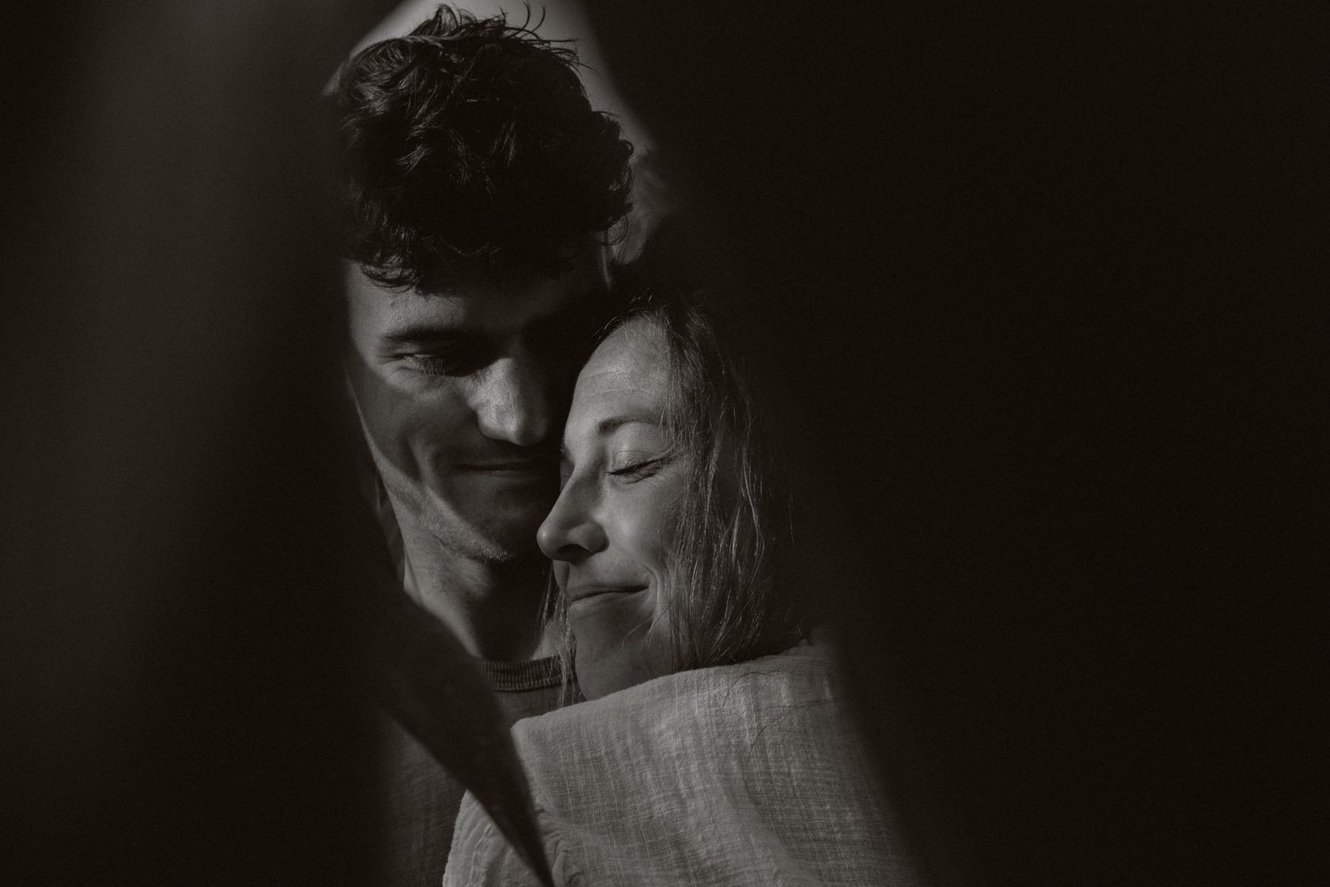 Black and white photo of a smiling couple embracing tenderly in soft shadow.